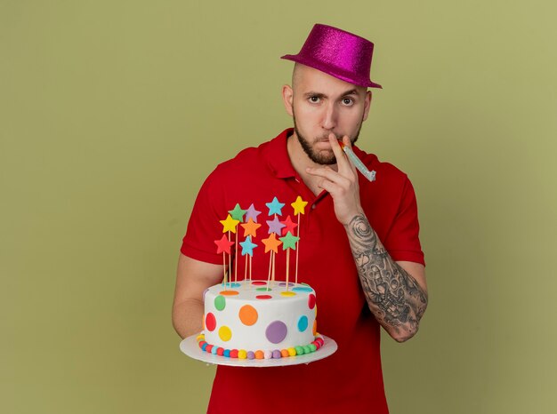 Zuversichtlich junger hübscher slawischer Party-Typ, der Partyhut hält, der Geburtstagstorte hält, die Kamera hält Party-Gebläse im Mund lokalisiert auf olivgrünem Hintergrund mit Kopienraum