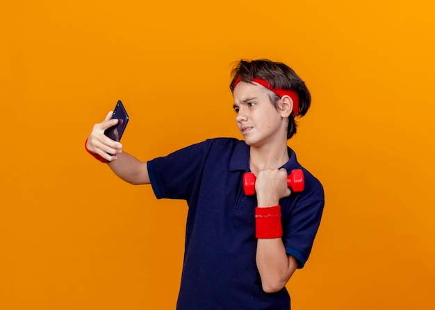 Zuversichtlich junger hübscher sportlicher Junge, der Stirnband und Armbänder mit Zahnspangen hält, die Hantel halten Selfie lokalisiert auf orange Wand