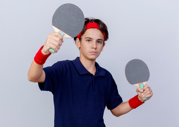 Zuversichtlich junger hübscher sportlicher Junge, der Stirnband und Armbänder mit Zahnspangen trägt, die Front halten und Ping-Pong-Schläger nach vorne strecken, lokalisiert auf weißer Wand