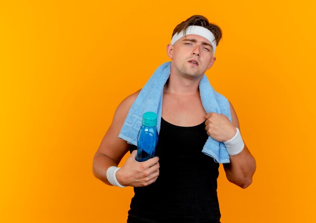 Zuversichtlich junger hübscher sportlicher Mann, der Stirnband und Armbänder mit Handtuch um den Hals hält Wasserflasche und Handtuch lokalisiert auf Orange mit Kopienraum