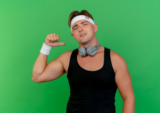 Zuversichtlich junger hübscher sportlicher Mann, der Stirnband und Armbänder mit Kopfhörern am Hals trägt, die auf sich lokalisiert auf Grün zeigen