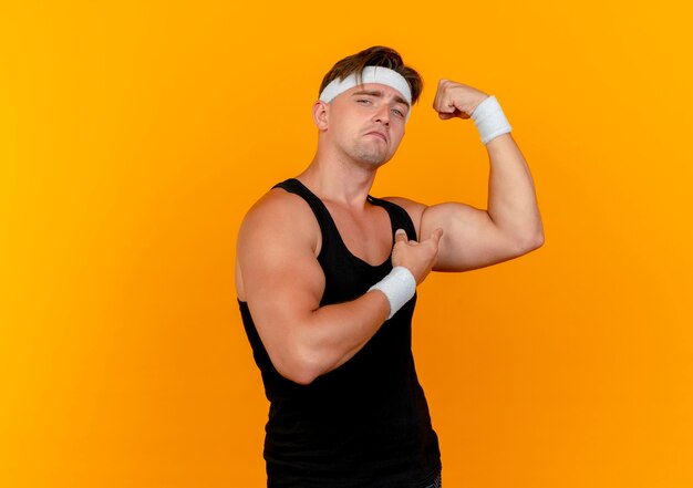 Zuversichtlich junger hübscher sportlicher Mann, der Stirnband und Armbänder trägt, die stark gestikulieren und auf seine Muskeln zeigen, die auf Orange mit Kopierraum isoliert sind