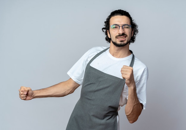 Zuversichtlich junger kaukasischer männlicher Friseur, der Brille und gewelltes Haarband im einheitlichen zwinkernden und geballten Fäusten trägt, lokalisiert auf weißem Hintergrund