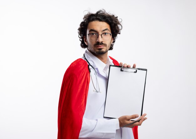 Zuversichtlich junger kaukasischer Superheldenmann in der optischen Brille, die Arztuniform mit rotem Umhang und mit Stethoskop um Hals hält, das Klemmbrett und Bleistift lokalisiert auf weißer Wand hält