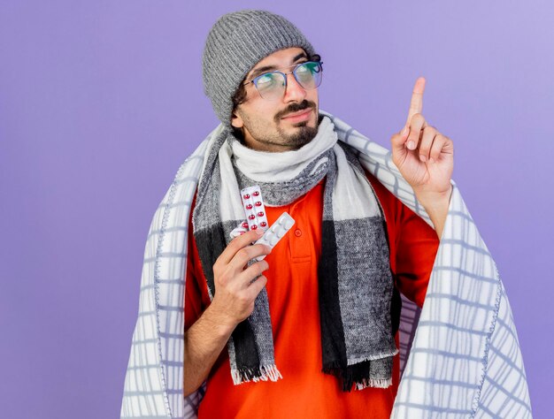 Kostenloses Foto zuversichtlich junger kranker mann, der brille wintermütze und schal in plaid hält, die packungen der medizinischen pillen hält, die lokal auf lila wand mit kopienraum suchen und zeigen