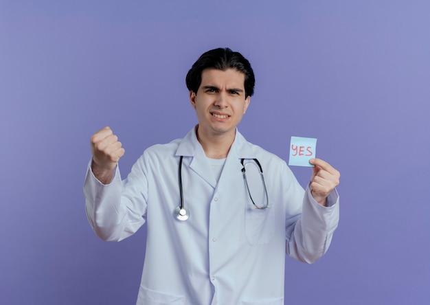 Zuversichtlich junger männlicher Arzt, der medizinisches Gewand und Stethoskop trägt, die ja Note zeigen, die starke Geste sein, die auf lila Wand mit Kopienraum isoliert wird