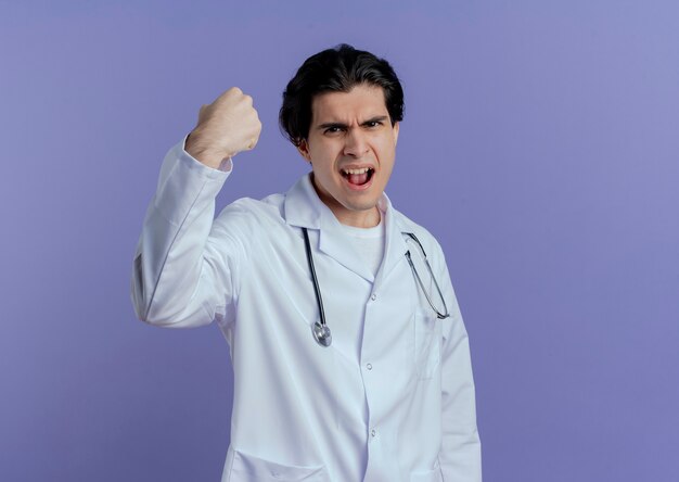 Zuversichtlich junger männlicher Arzt, der medizinisches Gewand und Stethoskop trägt und starke Geste tut, die auf lila Wand mit Kopienraum lokalisiert wird