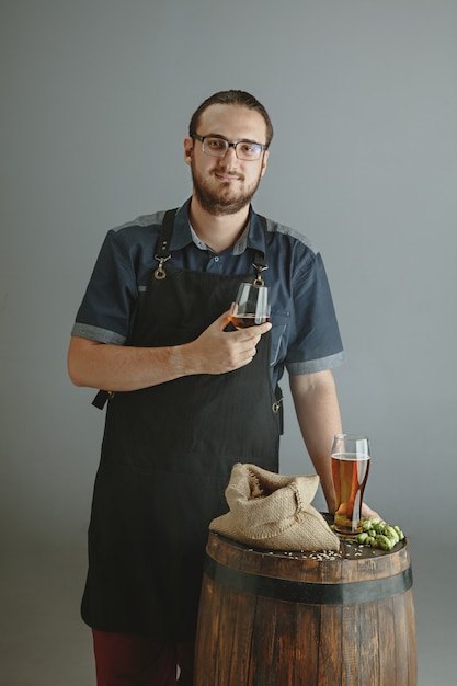 Zuversichtlich junger männlicher brauer mit selbst hergestelltem bier im glas auf holzfass auf grauem hintergrund.