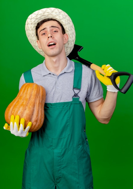 Zuversichtlich junger männlicher gärtner, der gartenhut und handschuhe trägt, hält kürbis und spaten auf schulter lokalisiert auf grünem hintergrund mit kopienraum