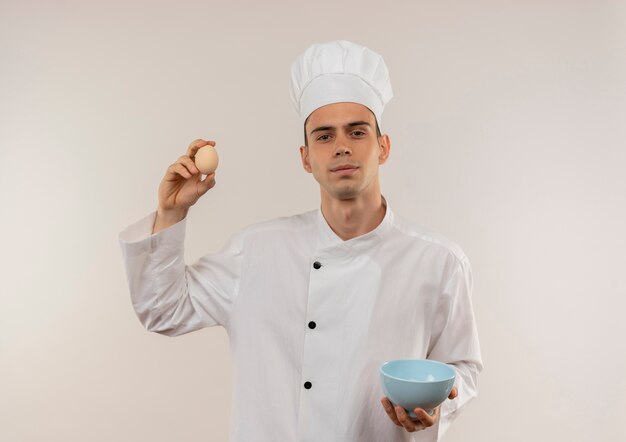 Zuversichtlich junger männlicher Koch, der Kochuniform hält Schüssel und Ei auf isolierter weißer Wand hält