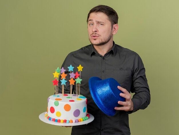 Zuversichtlich junger Party-Typ, der schwarzes Hemd hält Kuchen mit blauem Hut lokalisiert auf Olivgrün trägt