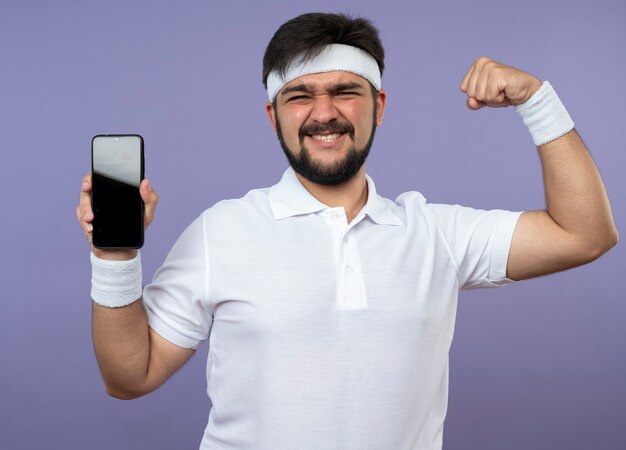 Zuversichtlich junger sportlicher Mann, der Stirnband und Armband hält Telefon hält und starke Geste zeigt, die auf Grün lokalisiert wird