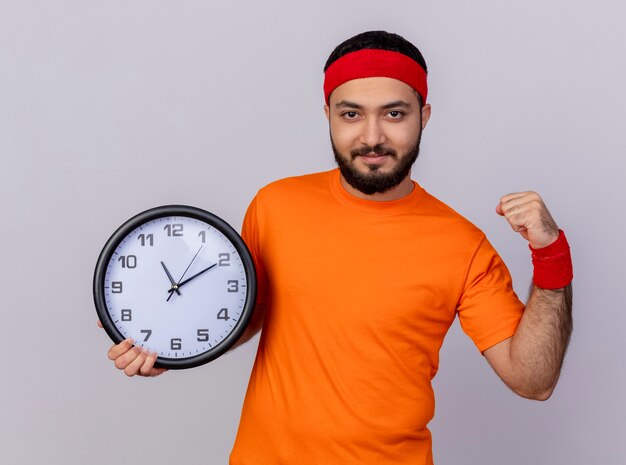 Zuversichtlich junger sportlicher Mann, der Stirnband und Armband hält Wanduhr zeigt starke Geste lokalisiert auf weißem Hintergrund