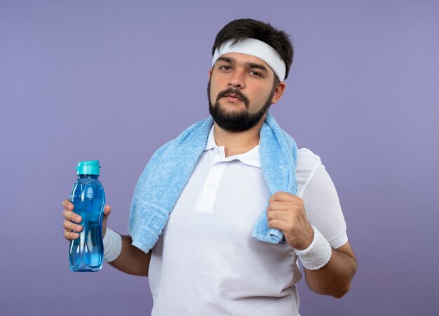 Zuversichtlich junger sportlicher Mann, der Stirnband und Armband mit Handtuch auf Schulter hält Wasserflasche trägt