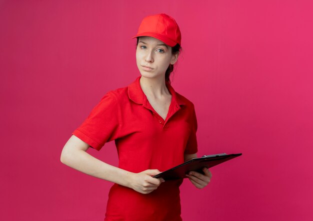 Zuversichtlich junges hübsches Liefermädchen in der roten Uniform und in der Kappe, die Zwischenablage hält, betrachtet Kamera lokal auf purpurrotem Hintergrund mit Kopienraum