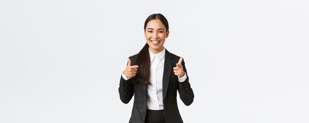 Zuversichtlich lächelnde asiatische Verkäuferin im schwarzen Anzug, die Daumen hoch zeigt, garantiert die Qualität des Produkts oder den besten Service. Verkaufsleiter, der versucht, etwas an den weißen Hintergrund des Kunden zu verkaufen