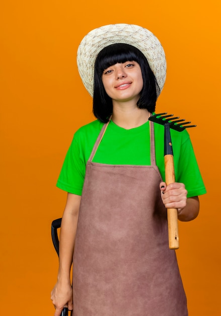 Zuversichtlich lächelnde junge Gärtnerin in Uniform mit Gartenhut hält Spaten und Rechen