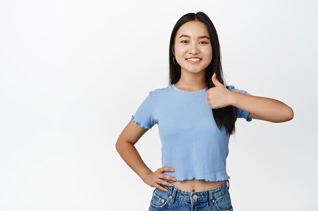 Zuversichtlich lächelndes asiatisches Mädchen zeigt Daumen nach oben, gibt Zustimmung, sagt ja und lobt das Stehen im T-Shirt auf weißem Hintergrund