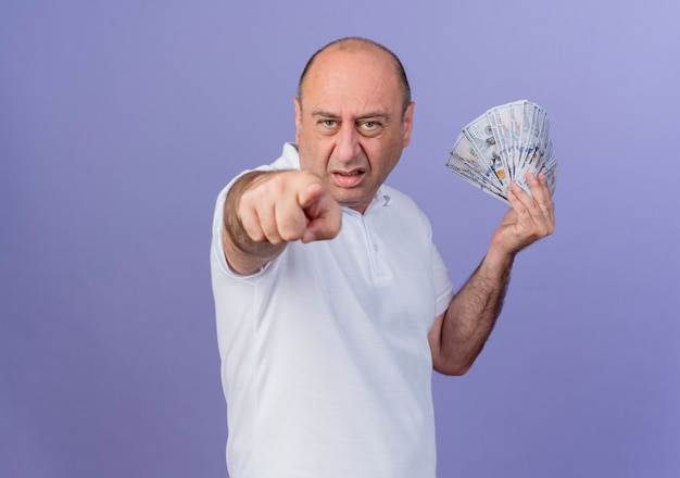 Zuversichtlich lässiger reifer Geschäftsmann, der Geld hält und auf Kamera lokalisiert auf lila Hintergrund mit Kopienraum zeigt