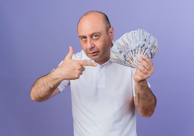 Zuversichtlich lässiger reifer Geschäftsmann, der Geld hält und auf purpurnen Hintergrund lokalisiert zeigt