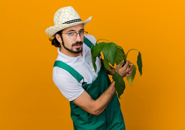Zuversichtlich männlicher Gärtner in optischen Gläsern, die Gartenhut tragen, hält Pflanze
