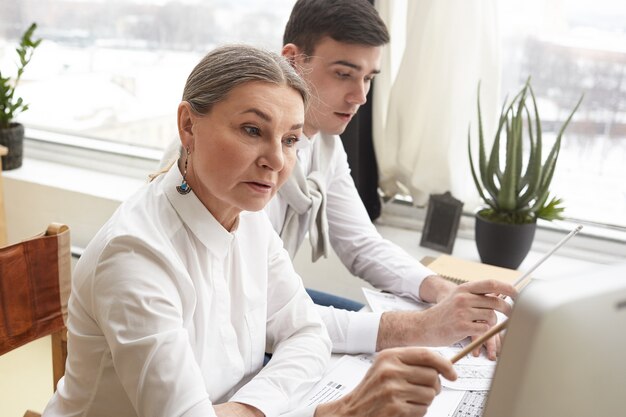 Zuversichtlich qualifizierte reife Ingenieurin, die Skizzen mit Computer und technischen Geräten macht und gemeinsam mit ihrem ehrgeizigen jungen Kollegen am Bauplan arbeitet. Talent und Kreativität