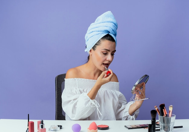 Zuversichtlich schönes Mädchen eingewickeltes Haartuch sitzt am Tisch mit Make-up-Werkzeugen, die Lippenstift halten und anwenden, der Spiegel lokalisiert auf lila Wand betrachtet