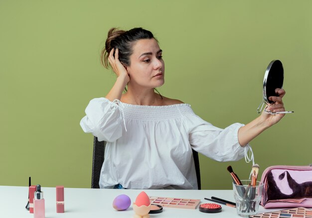 Zuversichtlich schönes Mädchen sitzt am Tisch mit Make-up-Werkzeugen legt Hand auf Kopf hält und schaut auf Spiegel isoliert auf grüner Wand