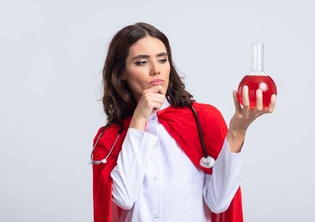 Zuversichtlich Superfrau in Arztuniform mit rotem Umhang und Stethoskop legt Hand auf Kinn und betrachtet rote chemische Flüssigkeit in Glaskolben isoliert auf weißer Wand