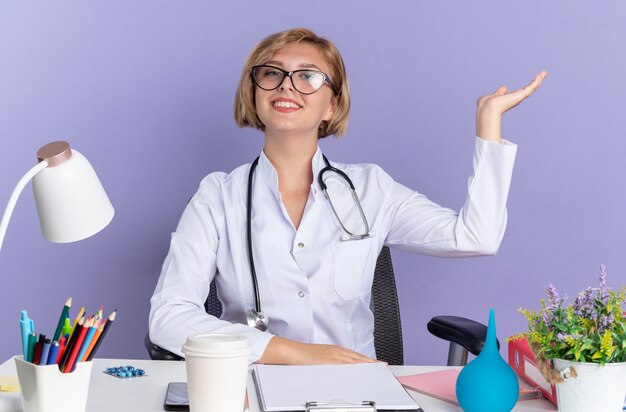 Zuversichtliche junge Ärztin, die ein medizinisches Gewand mit Stethoskop und Brille trägt, sitzt am Tisch mit medizinischen Werkzeugpunkten mit der Hand an der Seite isoliert auf blauer Wand mit Kopierraum