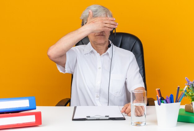 Zuversichtliche kaukasische Callcenter-Betreiberin auf Kopfhörern, die am Schreibtisch mit Bürowerkzeugen sitzen, die ihre Hand vor den Augen halten