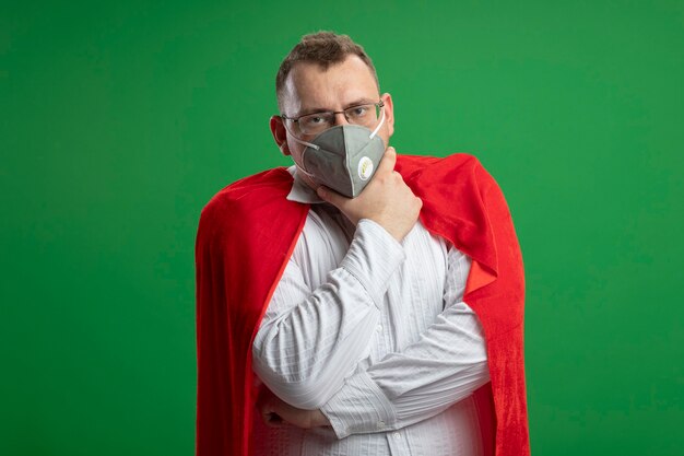 Zuversichtlicher erwachsener Superheldenmann im roten Umhang, der Brille und Schutzmaske trägt, die das vordere berührende Kinn lokalisiert auf grüner Wand betrachten