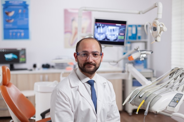 Zuversichtlicher Zahnarzt im Stomatologie-Kabinett mit orangefarbener Ausrüstung, die eine Zahnuniform trägt. Facharzt für Mundhygiene mit Laborkittel mit Blick auf die Kamera in der Zahnarztpraxis.