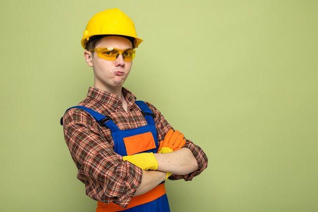 Zuversichtliches Überqueren der Hände junger männlicher Baumeister in Uniform und Handschuhen mit Brille