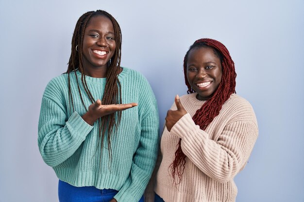 Zwei afrikanische Frauen, die vor blauem Hintergrund stehen, zeigen die Handfläche und machen eine OK-Geste mit Daumen nach oben, lächeln glücklich und fröhlich