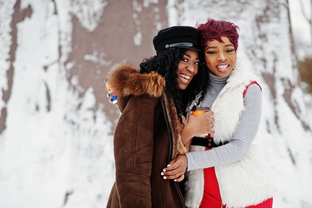 Zwei afroamerikanische Frauen in Schaffell und Pelzmantel posierten am Wintertag vor schneebedecktem Hintergrund mit Tassen Kaffee