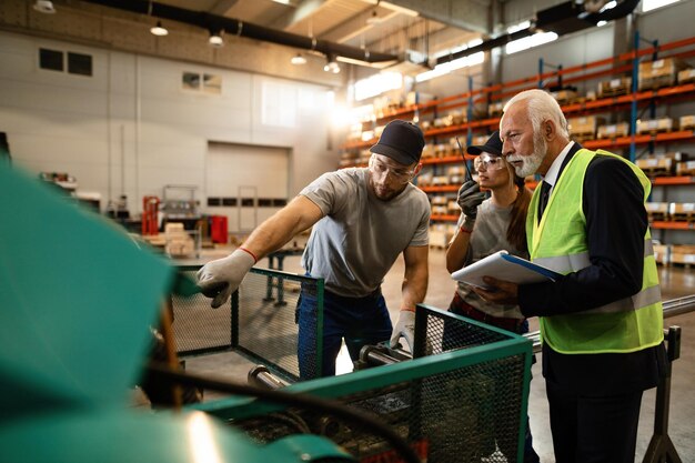 Zwei Arbeiter zeigen dem Qualitätskontrollinspektor eine Industriemaschine, die sie im Auslieferungslager verwenden