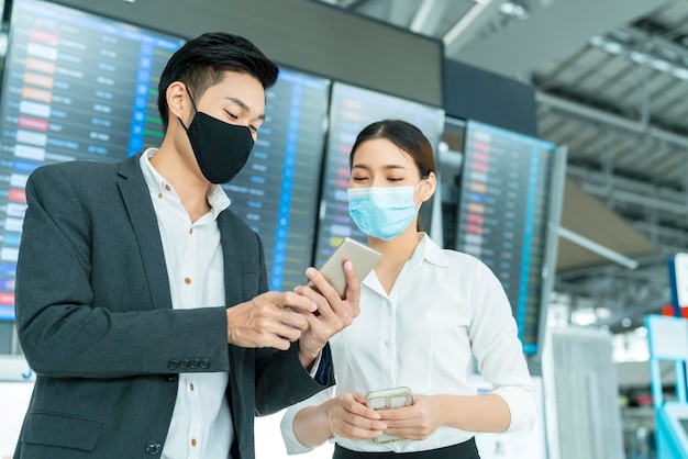 Zwei asiatische geschäftsleute und geschäftsfrauen mit gesichtsmaskenschutz im gespräch am internationalen flughafenterminal in der nähe des geschäftskonzepts der informationstafel