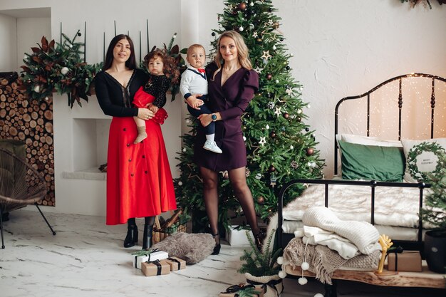 Zwei attraktive kaukasische Frauen mit ihren hübschen Kindern nahe dem Weihnachtsbaum zu Hause