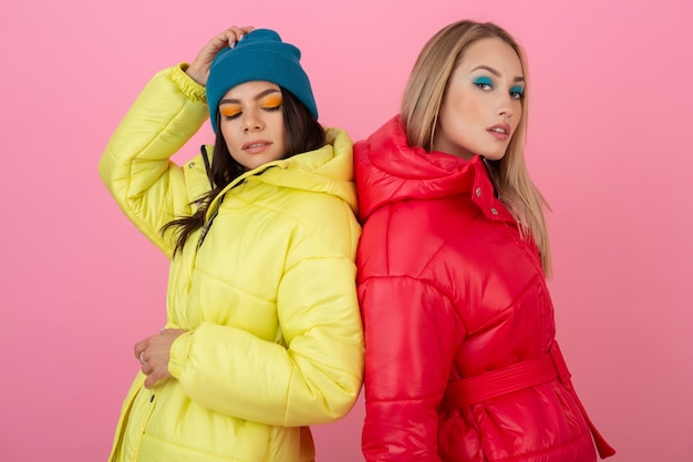 Zwei attraktive Mädchen, die auf rosa Hintergrund in der bunten Winter-Daunenjacke der leuchtend roten und gelben Farbe aufwerfen