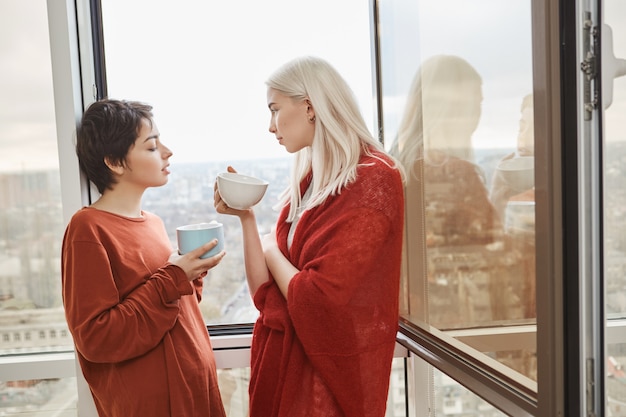 Zwei attraktive und sinnliche Freundinnen, die nahe beim geöffneten Fenster in roten Kleidern stehen, während sie Kaffee trinken