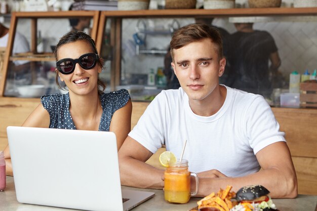 Zwei beste Freunde, die während des Mittagessens einen Laptop benutzen, in einem gemütlichen Café sitzen und mit einem glücklichen Lächeln schauen. Studenten, die online auf einem Notebook-PC lernen