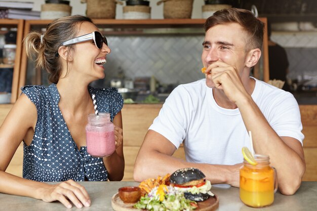 Zwei beste Freunde, die zusammen Spaß haben und lachen, während sie im Café zu Mittag essen. Attraktive Frau, die Glas des rosa Smoothie hält und lebhafte Unterhaltung mit ihrem hübschen Freund genießt