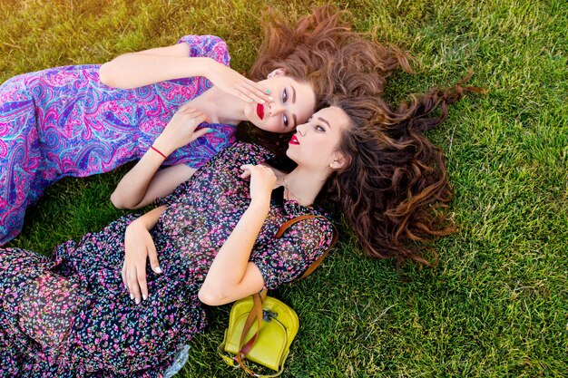 zwei beste Freunde in buntem Boho-Kleid und lockigem Haar, die auf dem grünen Gras liegen