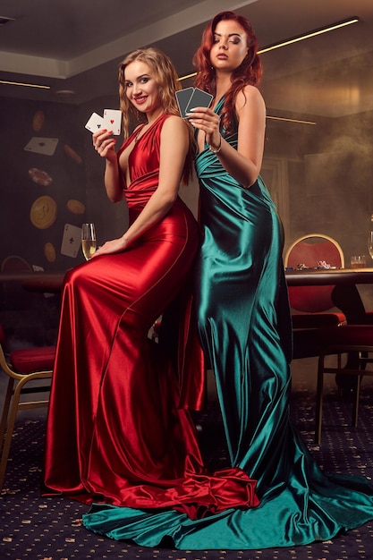 Zwei bezaubernde frauen in langen satinkleidern mit einigen spielkarten in der hand posieren an einem tisch in einem luxuskasino. leidenschaft, karten, chips, alkohol, gewinn, glücksspiel - es ist eine weibliche enterta
