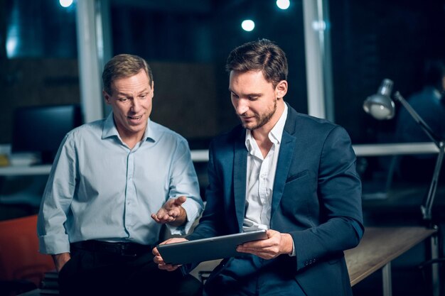 Zwei Büroangestellte mit Tablet im Büro spät in der Nacht. Junge Geschäftsleute unterhalten sich, während sie sich eine Präsentation auf einem digitalen Touchscreen-Tablet ansehen.