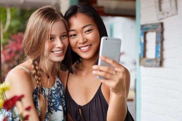 Zwei entzückende fröhliche asiatische und kaukasische junge Freundinnen machen Selfie auf modernem Handy, genießen Urlaub, reisen ins Ausland