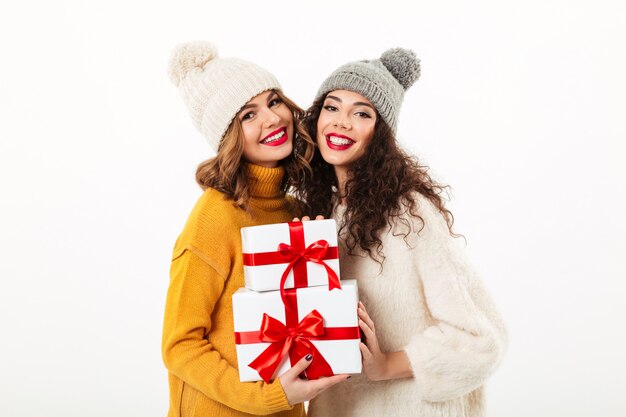 Zwei erfreute Mädchen in den Strickjacken und in Hüten, die beim Halten von Geschenken über weißer Wand zusammen stehen