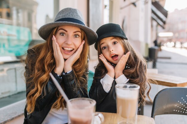 Zwei erstaunliche Mädchen in den trendigen Hüten, die mit lustigem Gesichtsausdruck während des Mittagessens im Straßenrestaurant am sonnigen Tag aufwerfen.