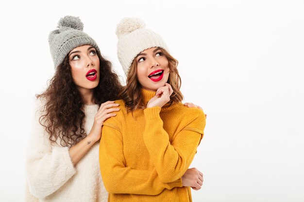 Zwei faszinierte Mädchen in den Strickjacken und in Hüten, die zusammen beim Schauen weg über weißer Wand stehen